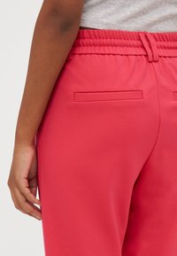 Gros plan d'une personne portant un pantalon rose vif avec taille élastique et poches arrière passepoilées, main détendue à ses côtés.