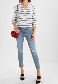 Weiß-blau gestreifte Bluse mit V-Ausschnitt, kombiniert mit hellblauen, zerrissenen Jeans und schwarzen Absatzsandalen. Eine rote Umhängetasche bringt Farbe ins Spiel.