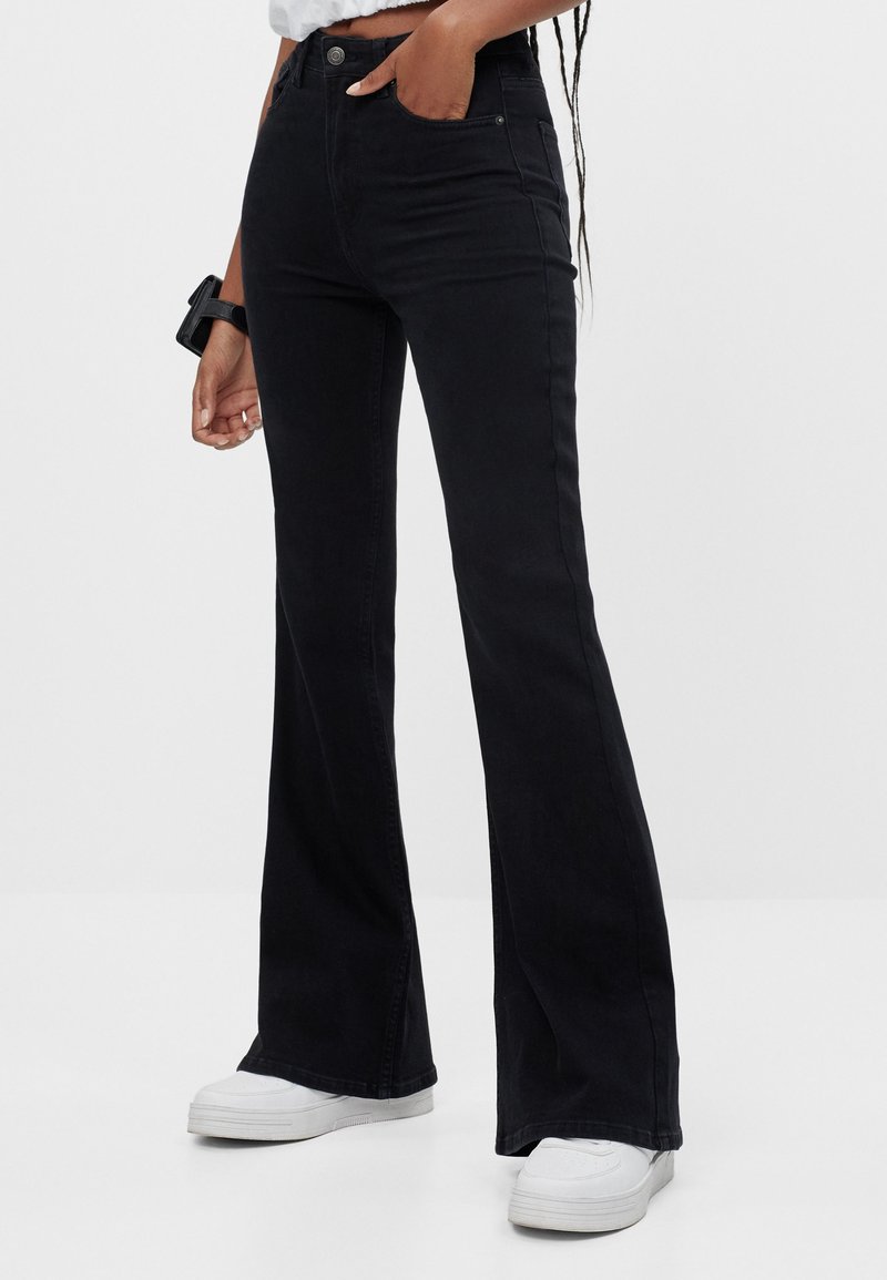 Jean en denim noir évasé à taille haute, avec poches avant et texture lisse. Assorti de baskets blanches sur un fond neutre.