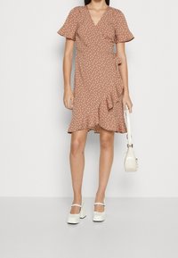 Vestido envolvente marrón de manga corta con lunares blancos, dobladillo con volantes y detalle de lazo. Llevado con tacones gruesos blancos y un bolso pequeño.