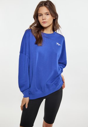 Blaues Sweatshirt aus weichem Stoff mit Rundhalsausschnitt, überschnittenen Schultern und einem kleinen weißen "Mrs."-Logo auf der Brust. Kombiniert mit schwarzen Shorts.