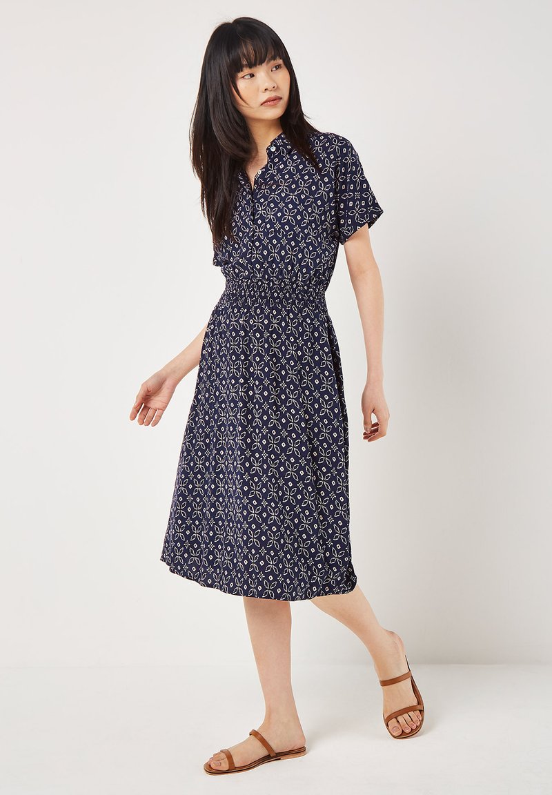 Navyblaues Kurzarmkleid mit weißem Blumenmuster, elastischem Bund, knielang, kombiniert mit braunen Sandalen. Glatte Stoffstruktur.