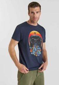Hombre con camiseta azul marino con un gráfico de un casco, gafas de sol, rosas azules y mariposas naranjas, combinada con pantalones verde oliva.
