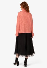 Maglione rosa lavorato a maglia con colletto alto, abbinato a una gonna in tulle plissettata nera. La modella indossa stivaletti neri con tacco a blocco.