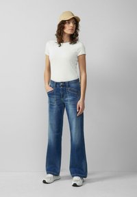 Frau mit beigem Hut, weißem Kurzarmhemd, weitem blauen Jeans mit vorderen Taschen und weißen Turnschuhen, die vor einem grauen Hintergrund steht.