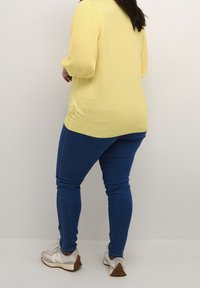 Gul, randig långärmad topp, blå slim-fit jeans och ljusgrå sneakers med bruna accenter, sedd bakifrån mot en neutral bakgrund.