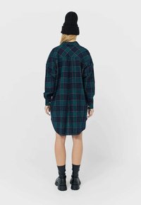 Skjortklänning med lång ärm i rutigt mönster i marinblå och gröna nyanser, med rundad nederkant och knapprygg, kombinerad med svarta knähöga stövlar.
