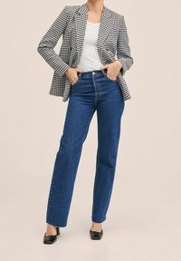 Blazer a motivi pied de poule, top bianco, jeans blue a gamba dritta a vita alta, tacchi neri, con uno sfondo neutro.