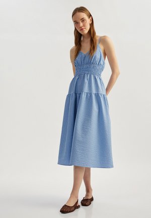 Sininen ruudullinen midi-mekko, jossa on säädettävät spaghetti-olkaimet, kootut rintakappaleet ja kerroksellinen helma. Yhdistetty ruskeisiin tasapohjaisiin kenkiin.