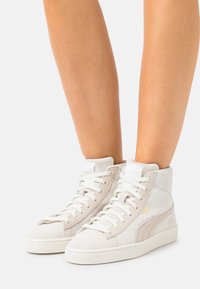 Białe i beżowe sneakersy za kostkę wykonane z zamszu, z sznurówkami, wyściełanym kołnierzem oraz teksturowaną gumową podeszwą z małym logo.