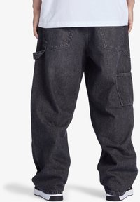 Henkilö, joka wearitsii löysiä tummanharmaita farkku cargo-housuja ja valkoisia lenkkareita, näkyy takaa vyötäröstä alas valkoista taustaa vasten.