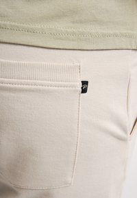 Détail de la poche arrière sur un pantalon beige avec une petite étiquette noire en tissu, associé à un haut vert olive clair dont l'ourlet est visible au-dessus de la taille.