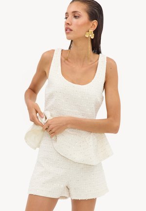 Femme portant un haut sans manches blanc texturé et un short assorti, tenant un détail de tissu, avec de grandes boucles d'oreilles florales dorées.