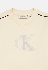 Crème-kleurige sweatshirt met ribgebreide kraag, voorzien van een verhoogd, geborduurd "CK"-logo in lichtgrijs. Horizontale zwarte strepen op de schouders.