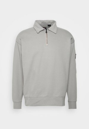 Persoon die een lichtgrijze sweater met kwart rits draagt, zwarte cargobroek met zijzakken en grijze lage sneakers, staand tegen een effen achtergrond.