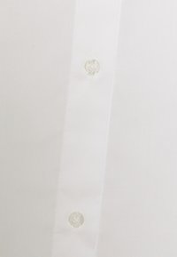 Tissu de chemise blanche avec gros plan sur une rangée verticale de deux boutons transparents cousus sur la patte de boutonnage.