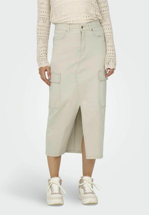Larga falda de mezclilla beige con una abertura frontal, bolsillos laterales y cierre de botones. Combinada con zapatillas blancas y un suéter de punto ligero.