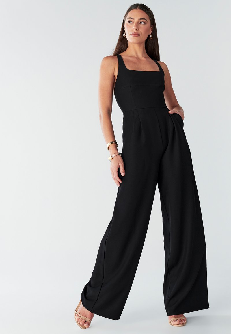 Zwarte jumpsuit met een vierkante halslijn, broek met wijde pijpen, naadloos ontwerp en zijzakken; model draagt nude sandalen met bandjes.