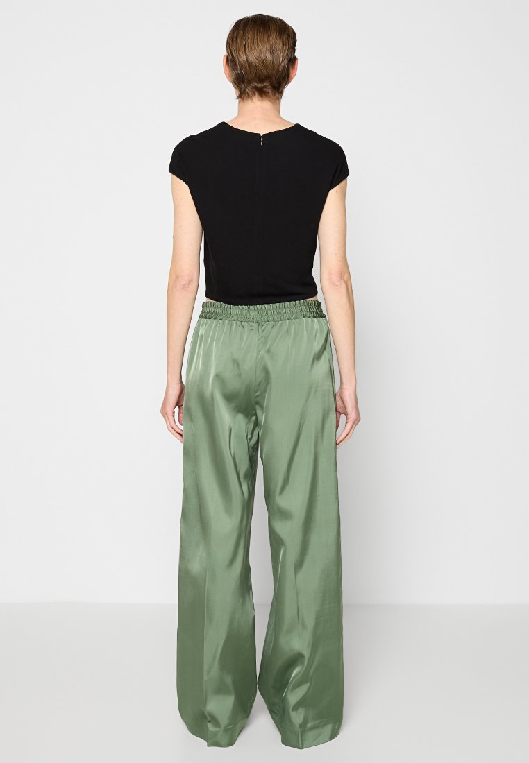 Schwarzes kurzärmeliges Oberteil mit einem Reißverschluss auf der Rückseite, kombiniert mit hoch taillierten, weit geschnittenen, glänzenden grünen Satinhosen. Minimalistisches Design.