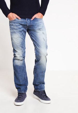 Man draagt vervaagde blauwe spijkerbroek en marineblauwe slip-on sneakers met witte zolen, handen in de zakken, staand tegen een witte achtergrond.