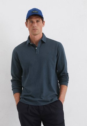 Long-sleeve navy polo shirt with a relaxed fit, three button placket, and subtle texture. Paired with dark trousers and a blue cap.