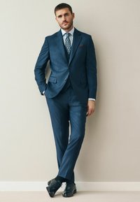 Dunkelblauer Anzug mit strukturiertem Finish, weißes Hemd, gemusterte Krawatte und schwarze Schuhe. Besonders hervorzuheben sind das Einstecktuch und die schlanke Passform.
