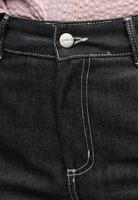 Μαύρο τζιν με κουμπιά και λευκή ραφή, φορεμένο με ροζ πουκάμισο με υφή, μπουφάν μέσα.