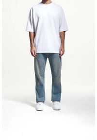 Valkoinen ylisuuri t-paita lyhyillä hihoilla, yhdistettynä vaaleansininisiin suoriin farkkuihin ja valkoisiin lenkkareihin neutraalilla taustalla.