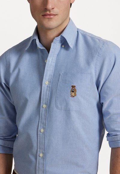 Chemise bleu clair à manches longues avec un tissu texturé, dotée d'une poche poitrine et d'un ours brodé portant un manteau.