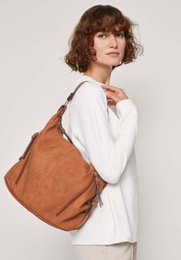 Bruin suède schoudertas met twee ritszakken aan de voorkant, een gebogen vorm en een verstelbare schouderriem, gedragen over een witte lange mouwen top.