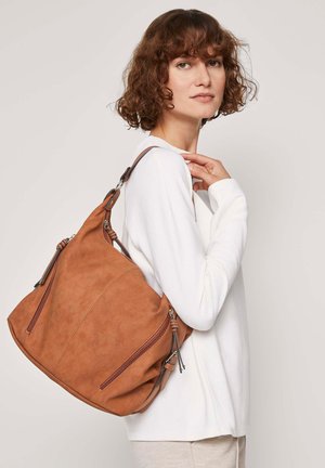 Braune Wildleder-Schultertasche mit zwei Reißverschluss-Fronttaschen, gebogener Form und verstellbarem Träger, getragen über einem weißen Langarmoberteil.