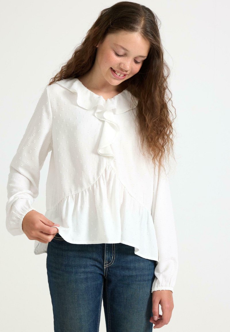 Tjej med långt lockigt hår som bär en vit blus med volang och blå jeans, leende och håller i blusens fåll.