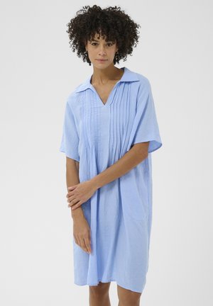 Žena s kučeravými vlasmi, oblečená v voľných svetlomodrých šatách po kolená s plisovaním a širokým golierom, stojaca so skríženými rukami pred bielym pozadím.