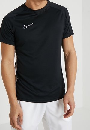 Homme portant un maillot de sport Nike noir à manches courtes et un short blanc, debout devant un fond clair uni.