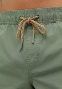 Shorts de bain vert clair avec taille à cordon de serrage. Le matériau semble lisse avec un léger éclat, présentant des coutures sur les côtés.