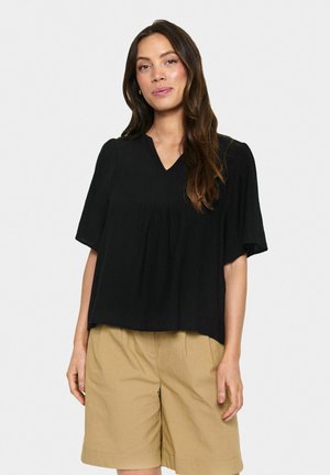Black short-sleeve top with a V-neck and gathered detailing, made from textured fabric, paired with beige shorts.