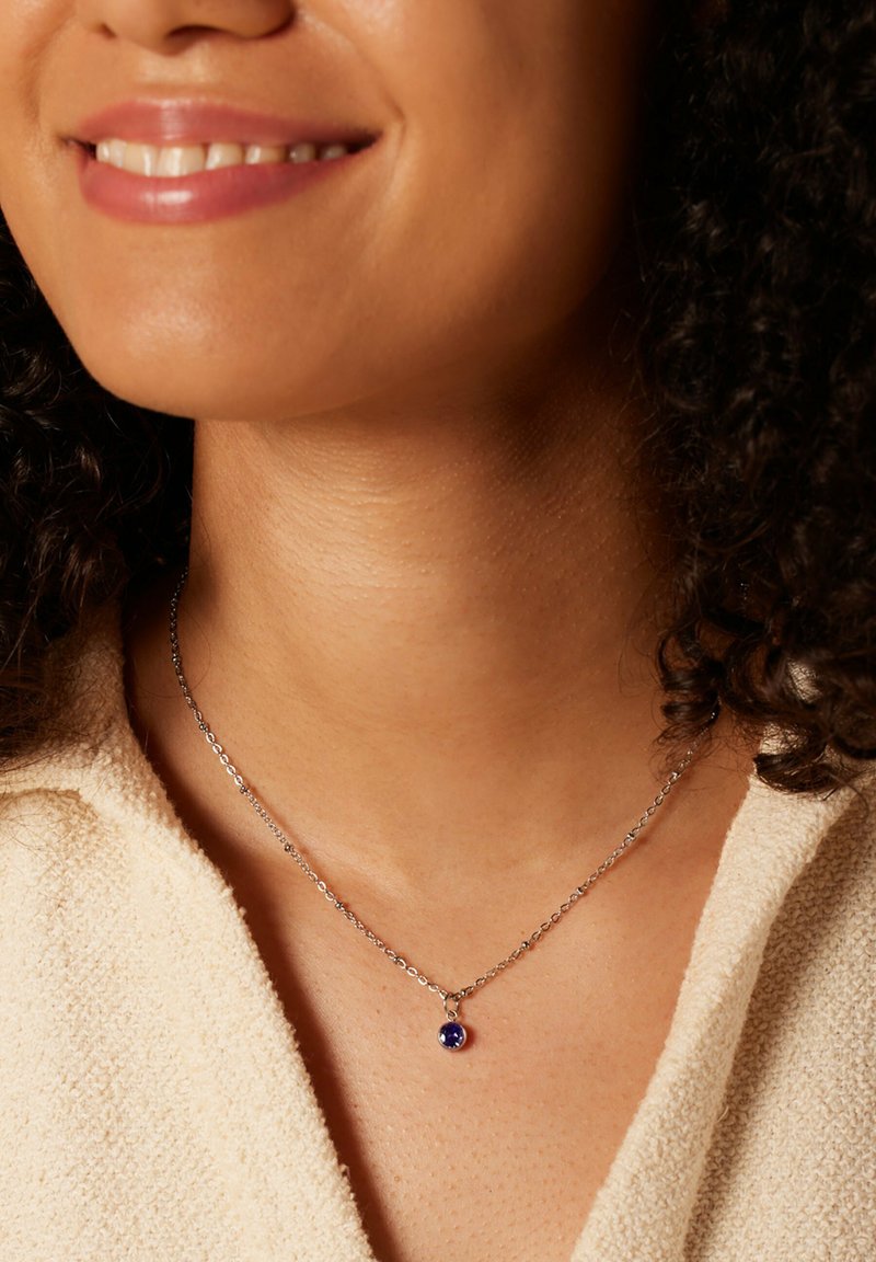 Zilveren ketting met een ronde paarse edelsteen hanger. De drager heeft krullend haar en de achtergrond toont een gestructureerde, lichtgekleurde stof.