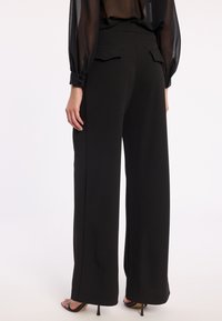 Zwarte wijde broek gemaakt van een soepele stof, met twee achterzakken en een hoge tailleband. Het model draagt zwarte hakken.