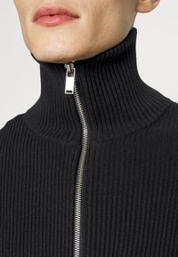 Maglione a collo alto nero a coste con una zip argentata, caratterizzato da un alto colletto e un tessuto testurizzato. Vista ravvicinata del colletto.