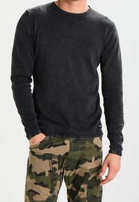 Camiseta negra de manga larga con cuello redondo, con una textura desgastada y costuras visibles, combinada con pantalones cargo de camuflaje.