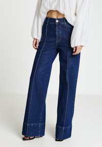 Jeans in denim a gamba larga a vita alta in blu scuro con cuciture a contrasto dorate, tasche grandi e orlo grezzo.