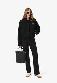 Veste noire courte avec une fermeture éclair frontale, un grand col et un logo brodé ; associée à un pantalon noir et un sac à main noir avec des poignées blanches.