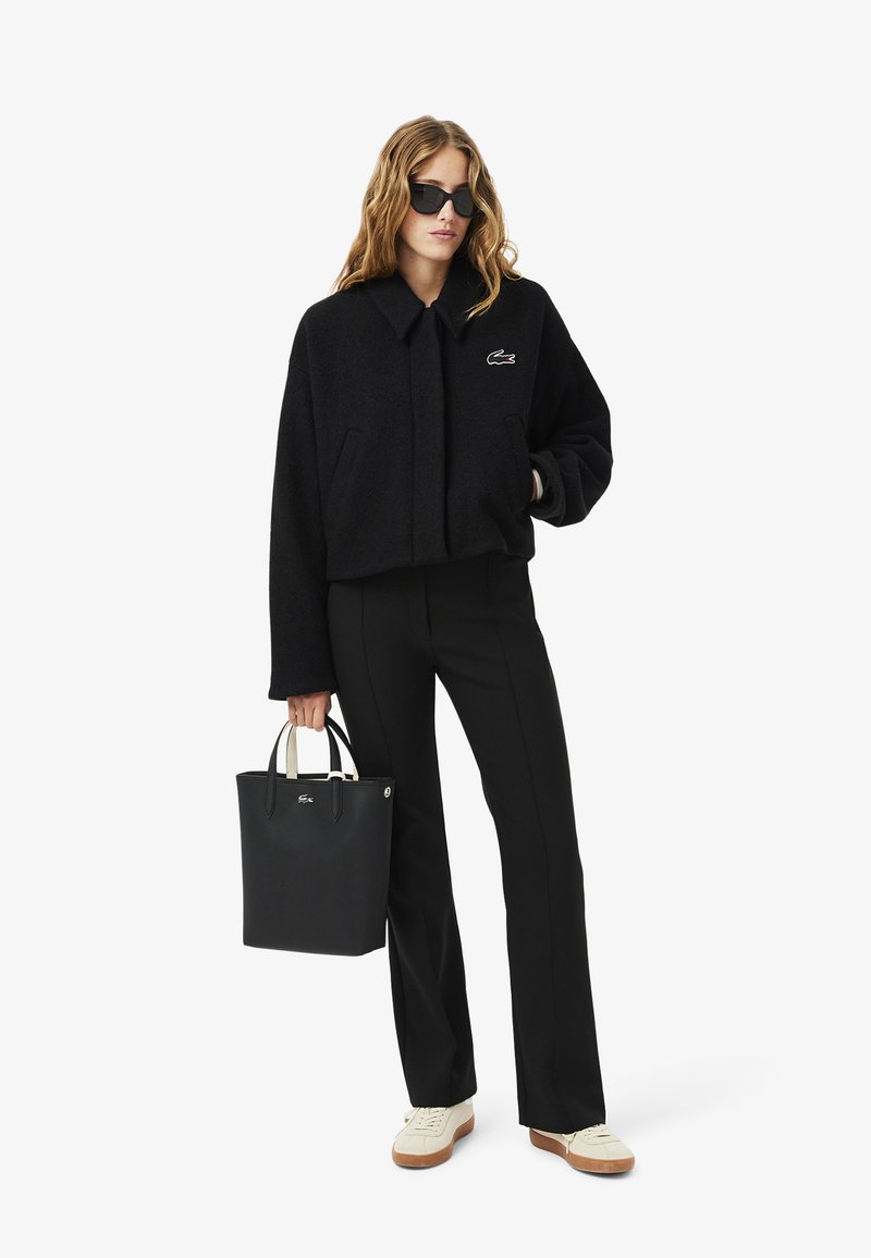 Veste noire courte avec une fermeture éclair frontale, un grand col et un logo brodé ; associée à un pantalon noir et un sac à main noir avec des poignées blanches.