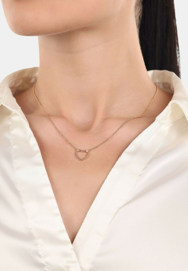 Collar de corazón en tono dorado con cuentas decorativas, llevado sobre una blusa de satén blanca. La cadena es fina, complementando el delicado diseño del colgante.