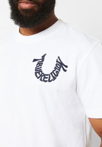 Camiseta de algodón blanca con cuello redondo, que presenta un logo bordado en color marino de "True Religion" en el lado izquierdo del pecho. Textura suave.