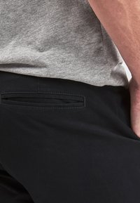 Schwarze Hosen mit glatter Textur, ausgestattet mit einer Gesäßtasche und sauberen Nähten. Kombiniert mit einem hellgrauen T-Shirt.