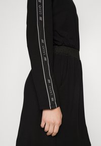 Svart långärmad klänning med ett texturerat bälte, vita och silverfärgade ränder längs ärmarna, samt sidfickor för funktionalitet.