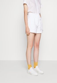 Hvide broderede shorts med bælte, parret med en hvid stribet kortærmet skjorte og hvide sneakers, suppleret med gule mønstrede sokker.