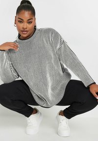 Maglione over-size a strisce bianche e nere con texture a coste e scollatura rotonda, abbinato a pantaloni neri aderenti e sneakers bianche.