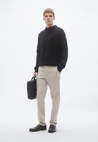 Sort strikket sweater, beige bukser og sorte loafers. Modellen står med en sort kuffert mod en hvid baggrund.